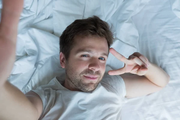 Красивый мужчина в белой рубашке делает селфи в постели — стоковое фото