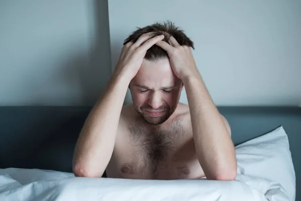 Занепокоєний чоловік сидить на ліжку з рукою на лобі. У нього проблеми з психічним здоров'ям і чути голоси в голові — стокове фото