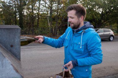 Mavi ceketli genç beyaz adam Almanya 'da bir geri dönüşüm konteynerine cam kavanoz atıyor.