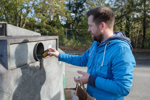 Młody biały mężczyzna w niebieskim płaszczu wyrzucający szklany słoik do pojemnika na recykling w Niemczech — Zdjęcie stockowe