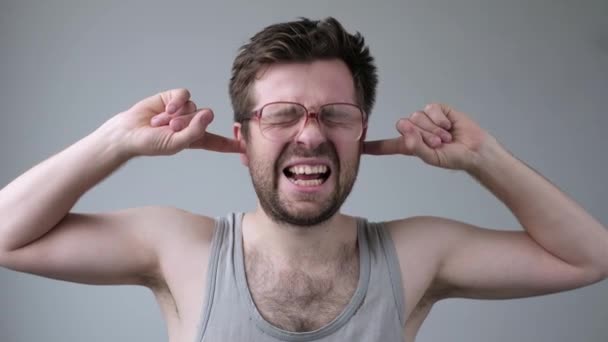 Jovem em óculos enormes ouvidos fechados com os dedos sendo irritado com barulho alto ou música — Vídeo de Stock