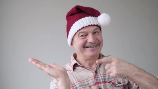 Confident content senior hispanic man in christmas hat pointing with forefinger aside — Stock Video