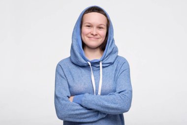 Mavi elbiseli ve kapüşonlu bir kadın gülümseyerek kameraya katlanmış elleriyle bakıyor.