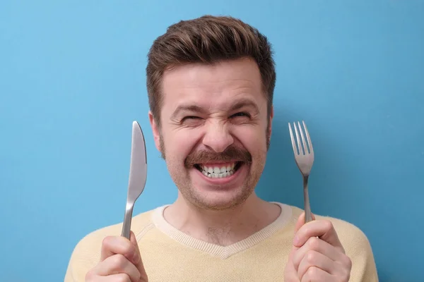 Un hombre loco y hambriento con bigote sostiene un cuchillo con un tenedor en las manos, sonríe. Me muero de hambre concepto . —  Fotos de Stock