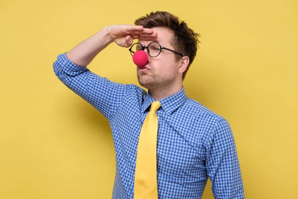 Χιλιετής άντρας με ρούχα κλόουν με κόκκινη μύτη και γυαλιά κοιτάζει μακριά στον κίτρινο τοίχο.. — Φωτογραφία Αρχείου