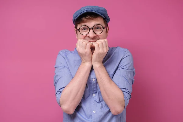 Junger lustiger Mann mit Brille und Hut beißt sich die Nägel an rosa Wand. — Stockfoto