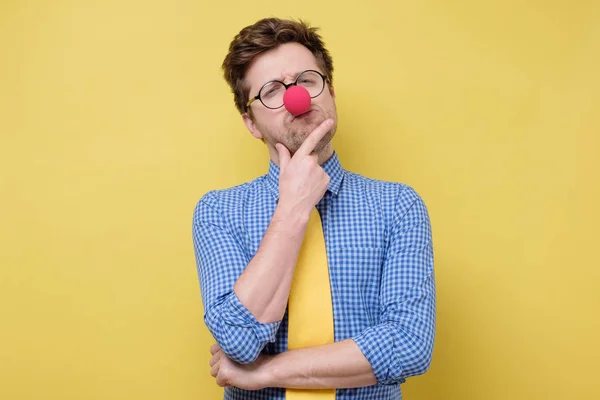 Funny caucasian man in red clown nose and yellow tie thinking with a hand in his chin