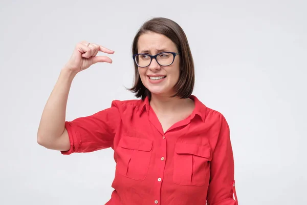Mulher madura em óculos e camisa vermelha mostrando algo pequeno e minúsculo com gesto e olhando chocado — Fotografia de Stock