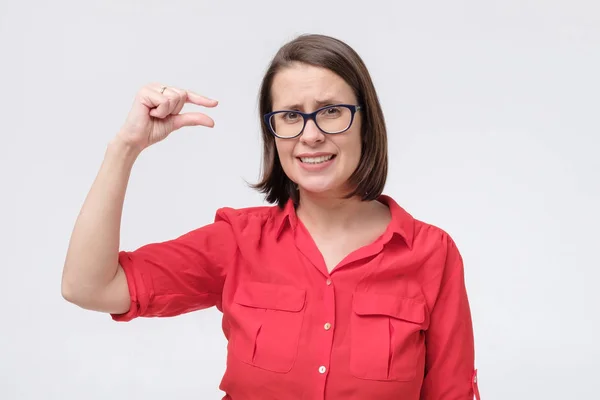 Mature woman in glasses and red shirt showing something small and tiny with gesture and looking shocked — Stock Photo, Image