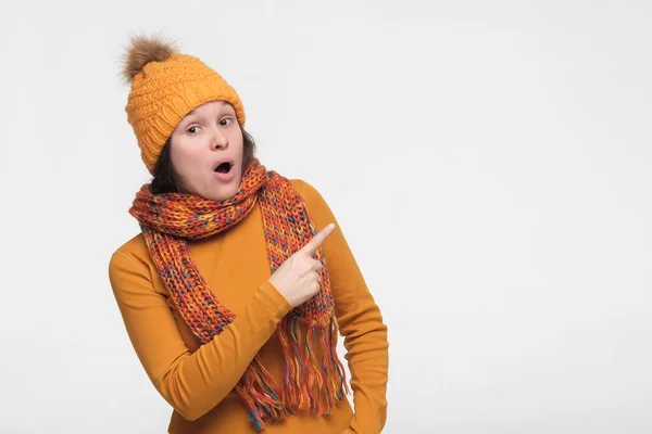 Attractive positive young female in winter clothes being surprised and pointing with forefingers away — Stock Photo, Image