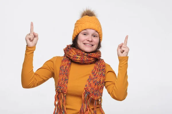 Atraente positivo jovem fêmea no chapéu de inverno e cachecol sorrindo alegremente e apontando com os dedos indicador de distância — Fotografia de Stock