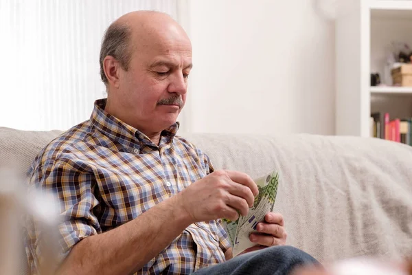 Senior man druk met het tellen van geld en rekeningen thuis, alleen thuis zitten. — Stockfoto