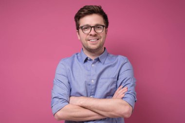 Kolları pembe arka planda çapraz duran gözlüklü gülümseyen genç adam.