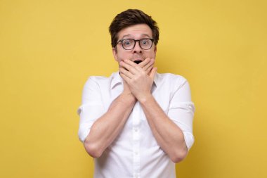 İmkanı yok. Beyaz gömlekli ve gözlüklü şok olmuş genç adam elleriyle ağzını kapatıyor ve sarı arka planda dikilirken haber veya satışlarla sersemlemiş bir şekilde kameraya bakıyor.