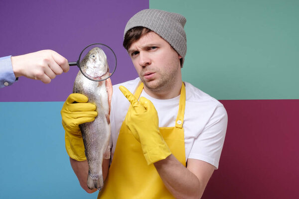 Young male fisher with yellow apron holding a big salmon checking its quality isolated on colored background. Selling fressh marine food.