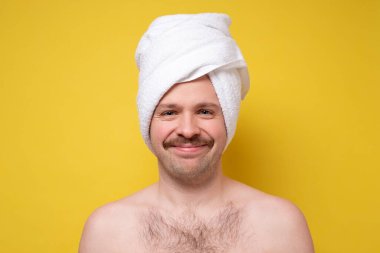 Bıyıklı ve kafasında beyaz havlu olan beyaz bir adam kameraya gülümsüyor. İnsan sağlığı için vücut ya da cilt bakımı. Sarı arkaplanda stüdyo çekimi.