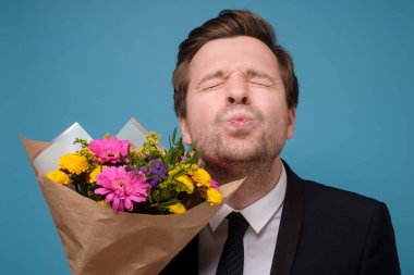 Komik, çekici, yetişkin bir adam öpüşmeyi beklerken çiçek buketi kıvrılmış dudaklarını tutuyor.