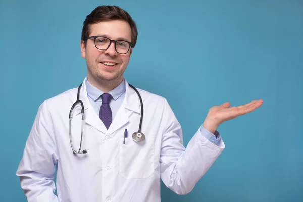 Joven médico caucásico con espectáculo de mano en copyspace sonriendo y mirando a la cámara — Foto de Stock