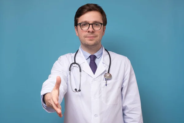 Young handsome caucasian man doctor in glasses giving handshake on blue wall — Stock Photo, Image