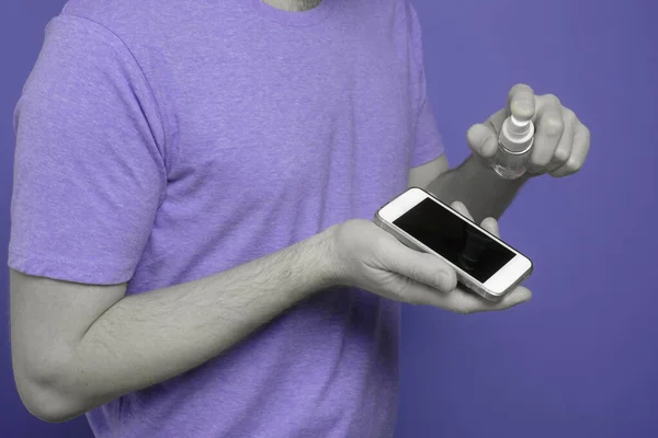 Hombre caucásico desinfectando su teléfono inteligente rociando un desinfectante de una botella . — Foto de Stock