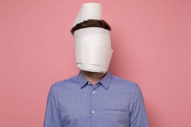 Mavi gömlekli bir adam yüzünü tıbbi maske olarak Coronavirus 'tan korumaya çalışan bir kağıda sarmış. Stüdyo resmi pembe duvarda..