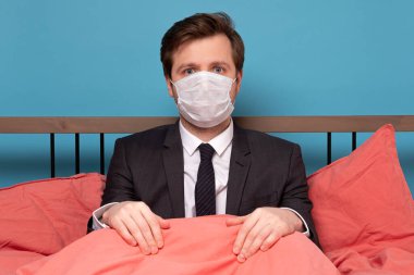 Yatakta oturan koruma maskesi takan bir iş adamı. Ev konseptinde kal. Virüs gribi ve COVID-19 humma karantinası.