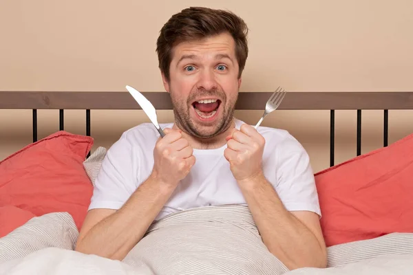 Young caucasian man is hungry holding fork and knife resting in bed. Stay home, wait for delivery and fellin happy. Studio shot