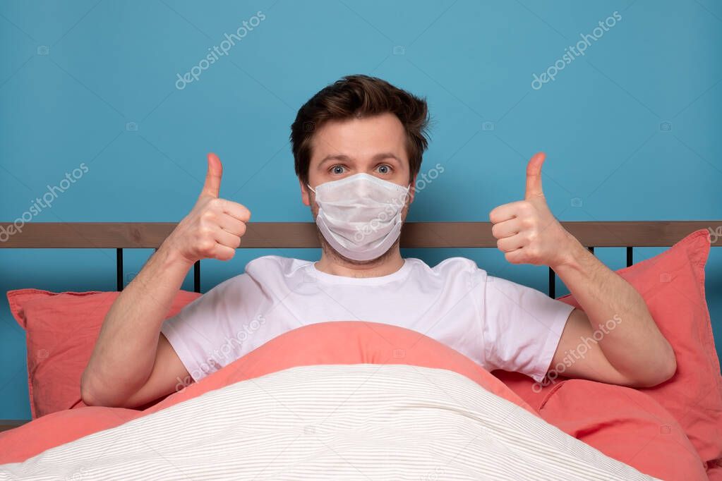 Young man wearing mask protection showing thumbs up staying home in quarantine. Studio shot.