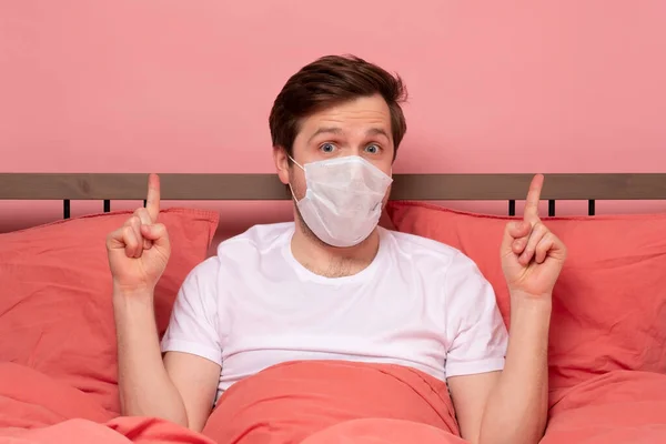 白人男性で医療マスクでベッドの上に安静時に何かを指しているピンクの背景 — ストック写真