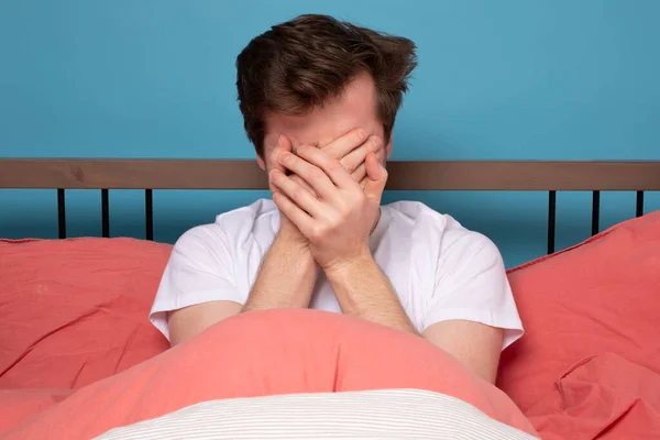 Scared young man covering face in stress sitting in bed. Quarantine or isolation concept. Negative facial human emotion.