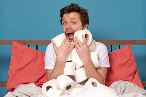 Excited happy caucasian man in bed hugging a pile of toilet paper isolated on blue background. Stay at home concept. Studio shot