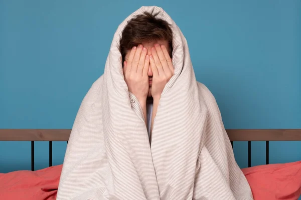 Scared young man covering face in stress sitting in bed. Quarantine or isolation concept.