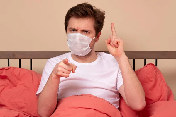 医療マスクの怒っている男はあなたが家のベッドに座って彼の指を指している 病気のときは家にいますか — ストック写真