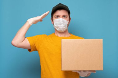 Tıbbi maskeli genç teslimatçı elinde bir kartpostal taşıyor ve mavi arka planda çalışmaya hazırlanıyor.