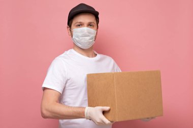 Tıbbi maskeli genç teslimatçı pembe arka planda izole edilmiş bir karton kutu taşıyor.