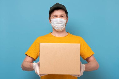 Maskeli genç teslimatçı mavi arka planda izole edilmiş bir karton kutu taşıyor. İnternetten karantina konsepti satın al.