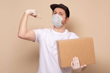 Tıbbi maskeli genç teslimatçı bej arka planda izole edilmiş pazıları gösteren bir karton kutu taşıyor.