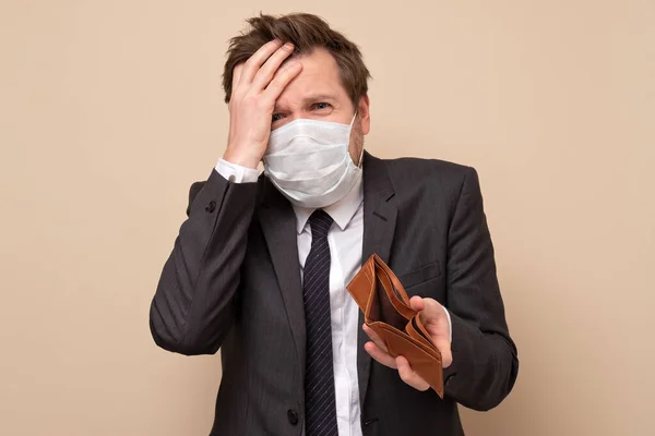 Poverty and absence of money. Unemployed sad caucasian man in medical mask and suit showing empty wallet, blue studio background
