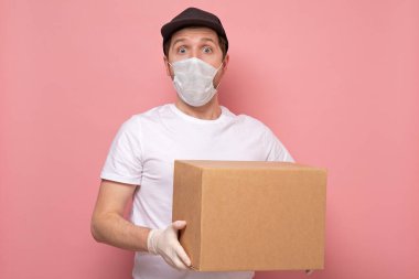 Tıbbi maskeli genç, sürpriz teslimatçı elinde beyaz arka planda izole edilmiş bir karton kutu taşıyor.
