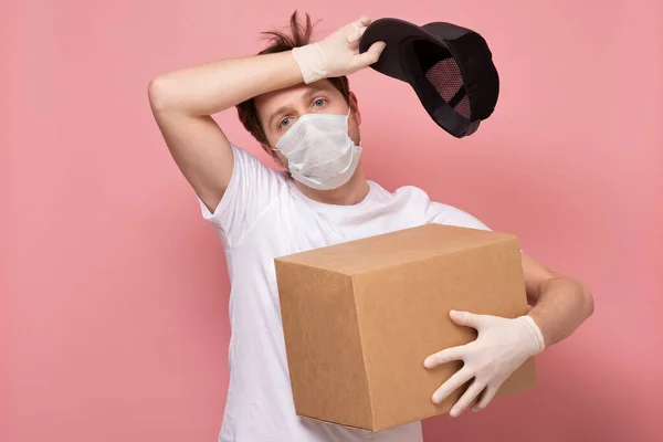 医療マスクを保持し 汗を拭く段ボール箱を運ぶ若い疲れと疲れ配達人 ピンクの背景に隔離されたまま仕事をしてる — ストック写真