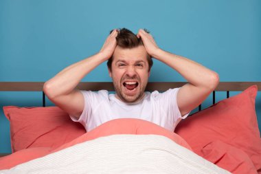 Stresli, yakışıklı genç bir adam yatakta yalnız başına oturuyor. Mutsuz ve yorgun hissedip başını tutuyor. Çalışma fotoğrafı.