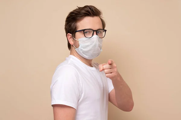 Caucasian young man in glasses with single use medical mask pointing you asking to stay home prevent infection, respiratory disease such as flu, 2019-nCoV. indoor studio shot isolated