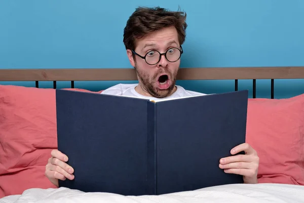Читання Захоплюючої Книги Сидить Ліжку Вдома Здивований Молодий Чоловік Окулярах — стокове фото