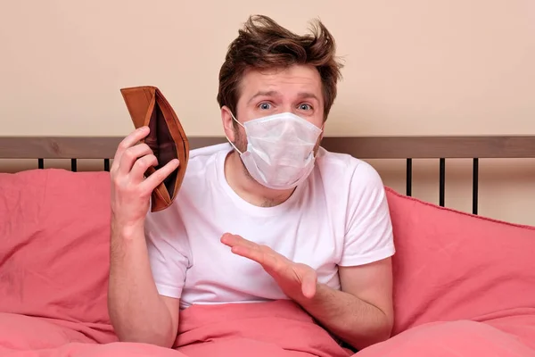 Poverty and absence of money. Unemployed sad caucasian man in medical mask sitting at home on quarantine or isolation because of covid showing empty wallet. He has no money for living.