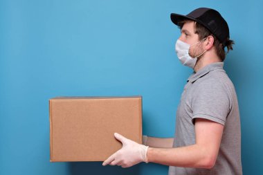 Tıbbi maske takan beyaz bir genç, kutusunu müşteriye veriyor. Teslimat konsepti yok. Stüdyo resmi mavi duvarda.