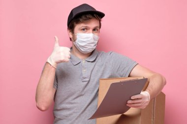 Tıbbi maskeli genç teslimatçı, elinde kart kutusu ya da pembe arka planda izole edilmiş bir paket olduğunu gösteriyor.