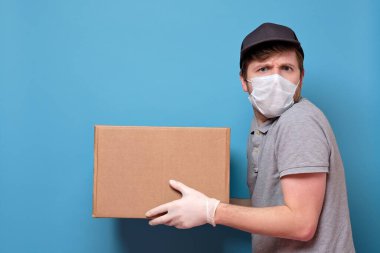 Tıbbi maske takan beyaz bir genç, kutusunu müşteriye veriyor. Teslimat konsepti yok. Stüdyo resmi mavi duvarda.