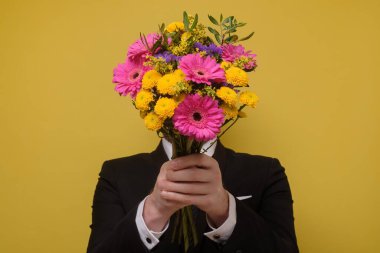 İsimsiz bir adam elinde çiçek buketiyle yüzünü sarı duvara gizliyor..