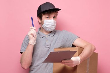 Tıbbi maskeli genç teslimatçı elinde bir karton kutu taşıyor ve pembe arka planda izole edilmiş kalem govong tavsiyesiyle işaret ediyor.