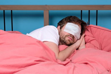 Tıbbi maske takan genç beyaz bir adam yatakta uyuyor. Stüdyo çekimi